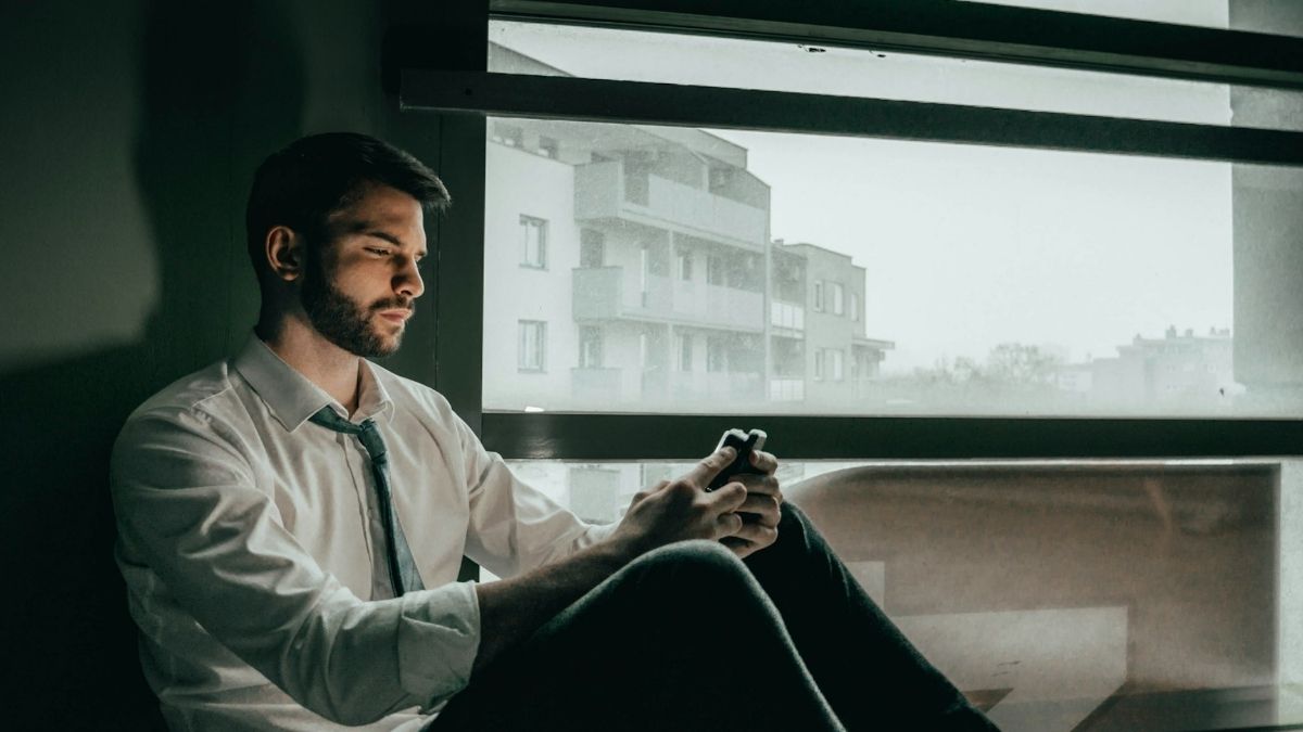 Nostalgija, muškarac sjedi gleda u mobitel i prisjeća se boljih vremena, vani pada kiša i tmurno je